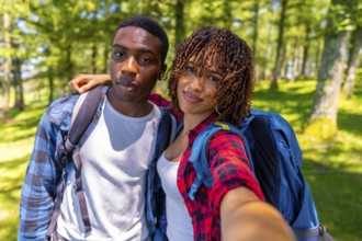 Two young hikers with backpacks smiling while taking a selfie in a sunlit forest, capturing the joy