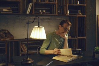 A middle-aged man in glasses sits at a dark wooden desk, deeply engaged in a book