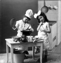 Two children cooking, 1920s, Two children playing in a kitchen, one in a cook's costume, in a black