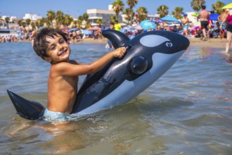 Smiling child having fun riding an inflatable orca toy in the shallow sea water by the beach on a