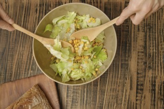 Hands are stirring a bowl of fresh salad featuring crisp lettuce and sweet corn. Nearby, a rustic