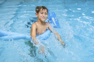 Smiling young boy splashing in a swimming pool, using a blue pool noodle. Joyful summer activity