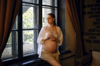 A pregnant woman stands by a large window in her home, holding a glass of water. She gazes outside,