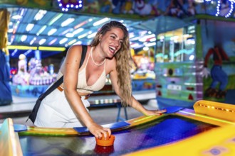 Cheerful young woman laughing and enjoying a lively game of air hockey at a vibrant amusement park,