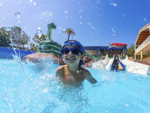 Young boy wearing blue sunglasses, joyfully splashing in a swimming pool at a vibrant water park,