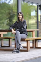 A young woman is seated at a wooden table outdoors, dressed in comfortable athletic wear. She