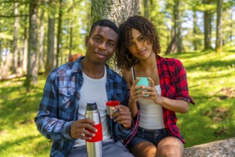 Young multi ethnic couple of hikers enjoying a hot drink from a thermos during a break in the woods