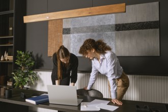 Two women discuss a project at a contemporary office desk, engaging with a laptop and scattered