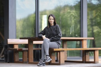 A young woman sits comfortably at a large wooden table placed outdoors. She wears a casual outfit