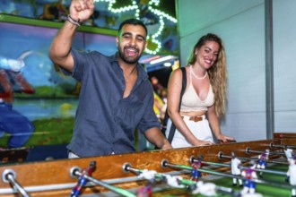 Young man and woman enjoying a foosball game at a funfair, celebrating a goal with excitement and