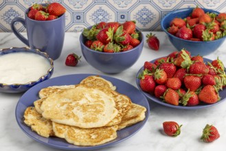 Pancakes on a plate, surrounded by bowls of strawberries and a bowl of yoghurt