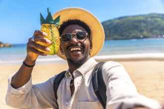 Young man wearing sunglasses and straw hat holding pineapple shaped cocktail while taking selfie on