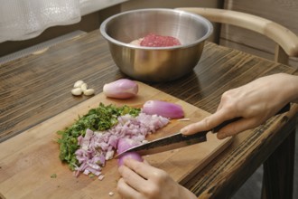 A person chops purple onions and fresh herbs on a wooden cutting board. A bowl of ground meat sits