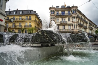 City fountain with running water in front of historic buildings on a cloudy day, Baden Baden,