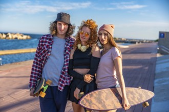 Three friends with skateboards relax by the seaside, enjoying the sunny weather and casual outdoor