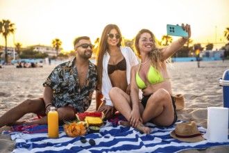 Three friends are enjoying a beach picnic at sunset, taking a selfie with fresh fruit and snacks