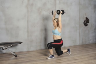 A woman is engaged in a strength training workout, lifting dumbbells overhead while in a lunge