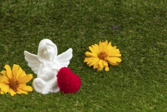 An angel figure with a red heart and yellow flowers on green grass