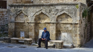 Man with blue hat sitting in front of a ruin-like facade with benches, fountain, Rhodes Old Town,