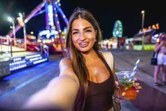 Happy young woman enjoying a vibrant night at an amusement park, taking a selfie while holding a