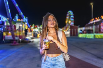 Young woman sips a beverage at a lively amusement park, illuminated by colorful lights, creating a