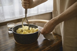 A person is mashing boiled potatoes in a pot. His hand grips a masher while holding a pot with