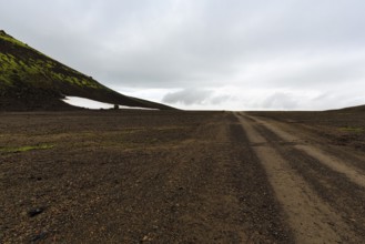 Gravel track, barren volcanic landscape with snowfields, mountain peaks with rain clouds,