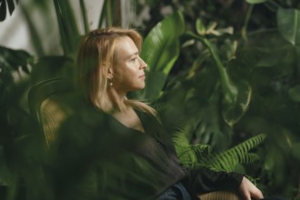 A woman sits quietly in a cozy chair within a vibrant indoor garden filled with lush plants and