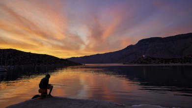 Angler sitting on the lakeshore during a colourful sunset against a mountainous backdrop, Holy