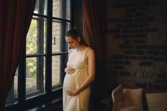 A pregnant woman stands peacefully by a large window in a cozy room, gently cradling her baby bump.