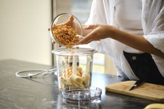 A person carefully adds sea buckthorn berries to a blender containing sliced bananas. The setting