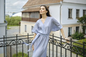 A woman stands on a balcony, wearing a flowing blue dress that drapes elegantly her slender body.
