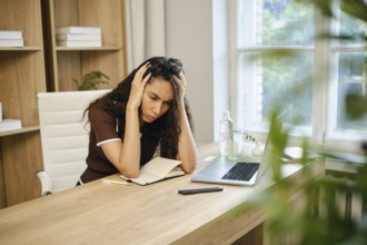 A young woman sits at her desk in a stylish home office, visibly stressed as she holds her head in
