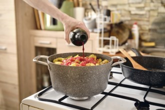 A person pours red wine into a pot filled with pork and vegetables making aromatic sauce
