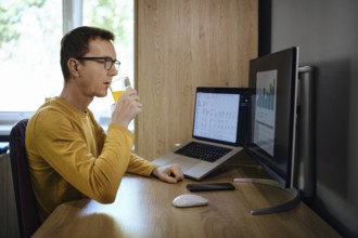 Individual is focused on work at a home office, sipping a drink while looking at two computer