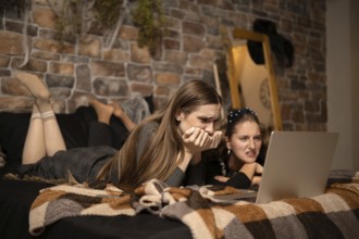 Two friends lie on a bed, deeply engaged as they watch an intense movie on a laptop. Their