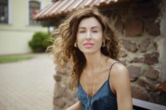 A woman with beautiful curly hair sits on a bench next to a rustic stone wall. She wears a stylish