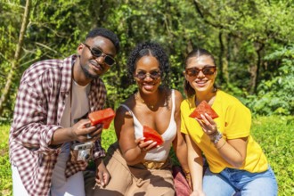 Three friends are sharing watermelon slices during a summer picnic in a lush park, enjoying the