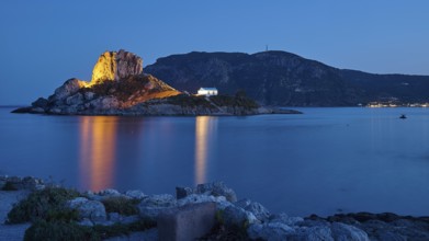 Rocks and buildings on an island, illuminated at night with reflecting water, Agios Stefanos Beach,
