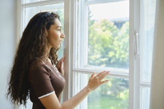 A young woman with long curly hair stands by a window, thoughtfully gazing outside. Sunlight