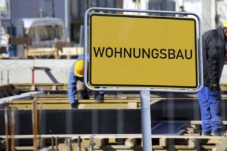 (Photomontage) Town sign with the inscription WOHNUNGSBAU in front of workers on a building site