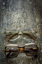 Two pairs of vintage eyeglasses rest on a weathered wooden surface, showcasing distinct styles. The