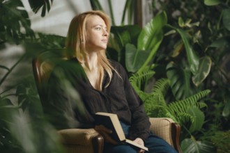 In a serene indoor garden, a woman sits comfortably in a chair holding a book. Soft light filters