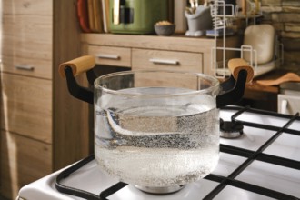 Water is boiling in a clear glass pot placed on a gas stove in a stylish kitchen. The scene
