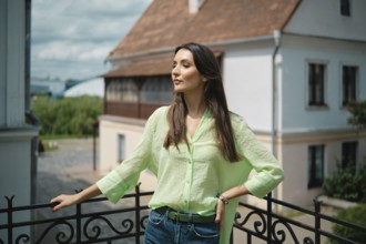 A young woman stands confidently on a balcony, wearing a light green blouse and blue jeans. She