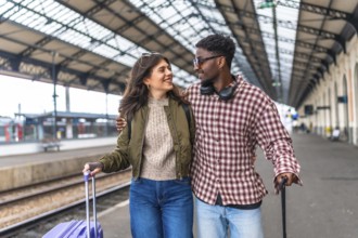 Young multi ethnic couple walking hand in hand with luggage on a bustling train station platform,