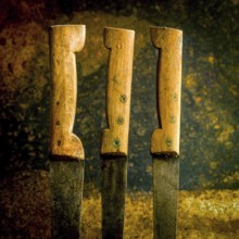 Three old, rusty kitchen knives with unevenly worn wooden handles rest against a textured backdrop.