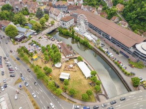 Park with people by a river, flanked by town buildings and a Ferris wheel, with heavy road traffic,
