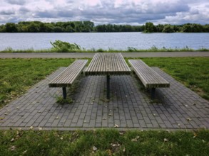 Empty picnic table with benches by a lake, surrounded by grass, under a cloudy sky in a park,