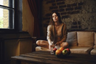 A pregnant woman is sitting on the couch near a window and reaching for a bottle of water on the